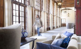 a woman is standing in a room with large windows and blue couches , holding a cup at Parador de Zamora