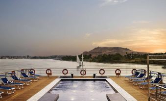 a large outdoor pool is surrounded by lounge chairs and a boat , with mountains in the background at The Oberoi Zahra, Luxury Nile Cruiser
