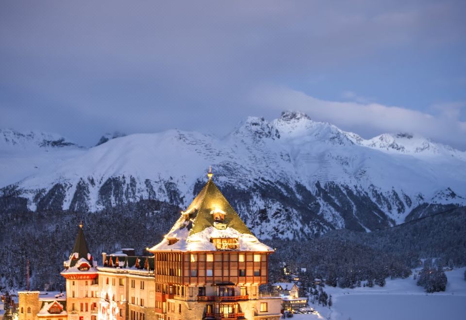 a large building with a golden dome is surrounded by snow - covered mountains at dusk , creating a picturesque winter scene at Badrutt's Palace Hotel St Moritz