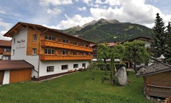 a large wooden building with multiple floors and balconies , situated in a grassy area surrounded by trees and mountains at Hotel Post