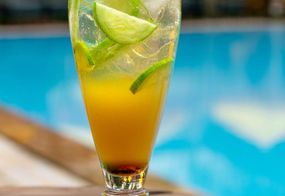 a tall glass filled with a yellow drink and garnished with lime slices is sitting on a wooden table near a pool at The Oberoi Grand Kolkata