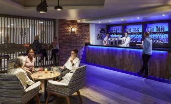 a group of people are sitting at tables in a bar with a brick wall and counter at Novotel Manchester West