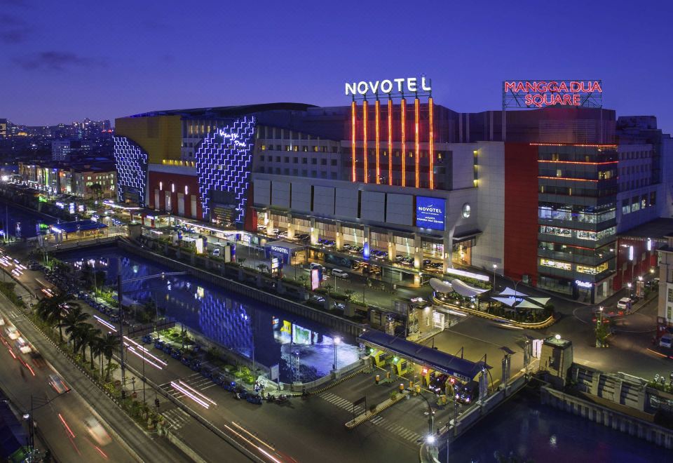 a busy city street at night , with multiple buildings and cars on the road , creating a bustling atmosphere at Novotel Jakarta Mangga Dua Square
