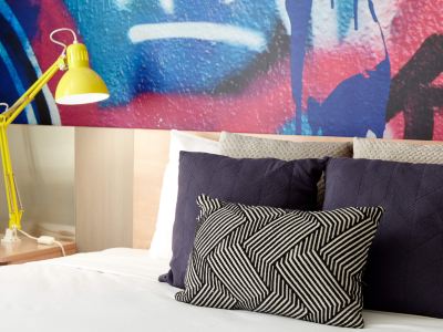 a bed with a black and white pillow on it , and a yellow lamp hanging above at Ibis Melbourne Hotel and Apartments