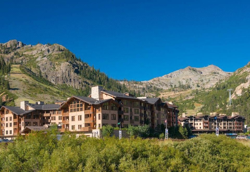 a large wooden building surrounded by trees and mountains , with a clear blue sky in the background at The Village at Palisades Tahoe