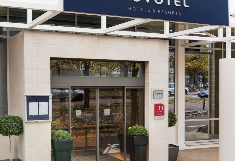 the entrance to the novotel hotel in europe , with the hotel 's name displayed above the door and two potted plants on either side of the at Novotel Château de Versailles