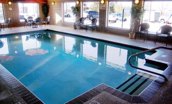 an indoor swimming pool with a hot tub , surrounded by lounge chairs and umbrellas , in a well - lit room at AmericInn by Wyndham Rogers