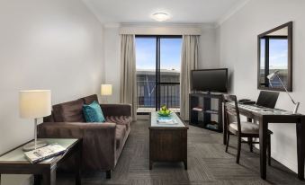 a living room with a brown couch , coffee table , and television near a window overlooking the city at Oaks Sydney North Ryde Suites