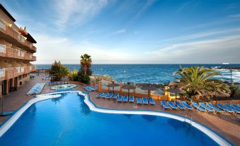 a large , blue swimming pool with a diving board and lounge chairs is surrounded by palm trees and the ocean at Elba Castillo San Jorge & Antigua Suite Hotel