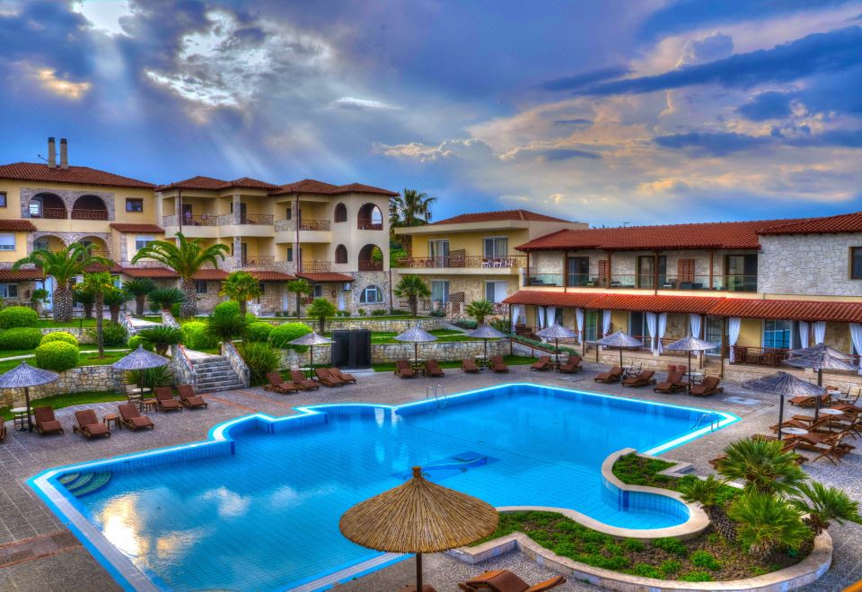 a large outdoor swimming pool surrounded by multiple buildings , with umbrellas and sun loungers placed around the pool area at Blue Bay Halkidiki