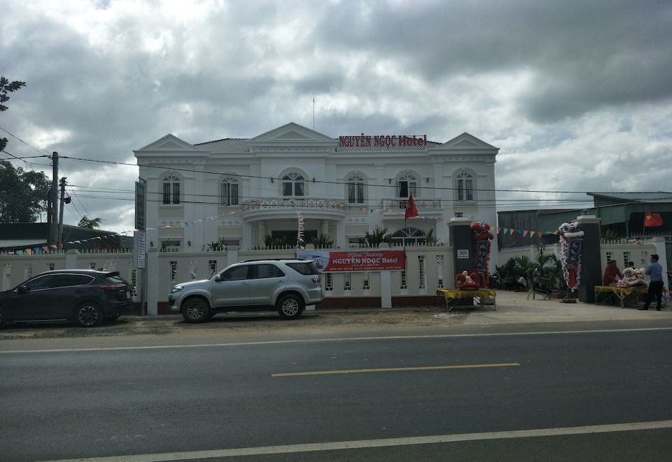 a white building with a red flag on the side is surrounded by cars and trees at Hotel Nguyen Ngoc