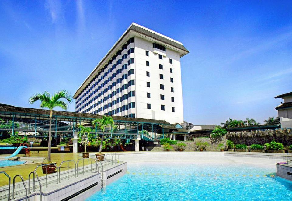 a large white hotel building surrounded by a swimming pool , with palm trees in the background at Horison Ultima Bandung