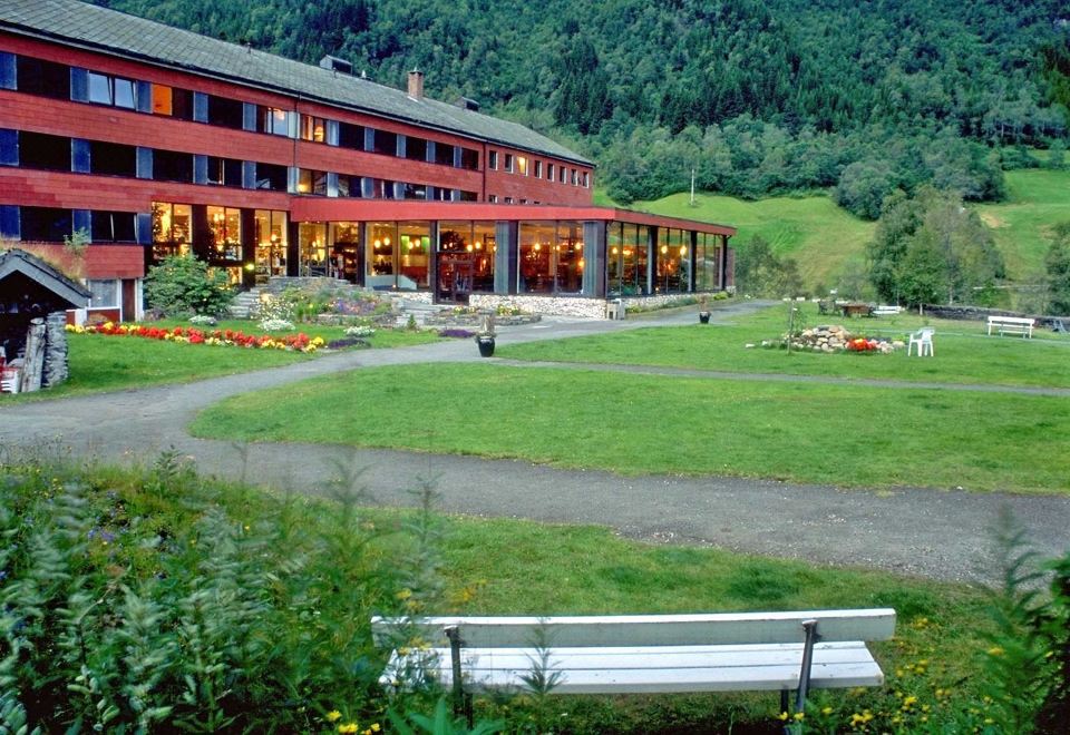 a large building with a red roof is surrounded by greenery and a white bench at Stalheim Hotel