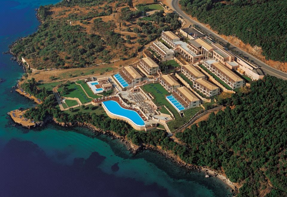 An aerial view displays a resort and the water surrounding it, featuring a prominent pool in the foreground at Ionian Blue Bungalows and Spa Resort