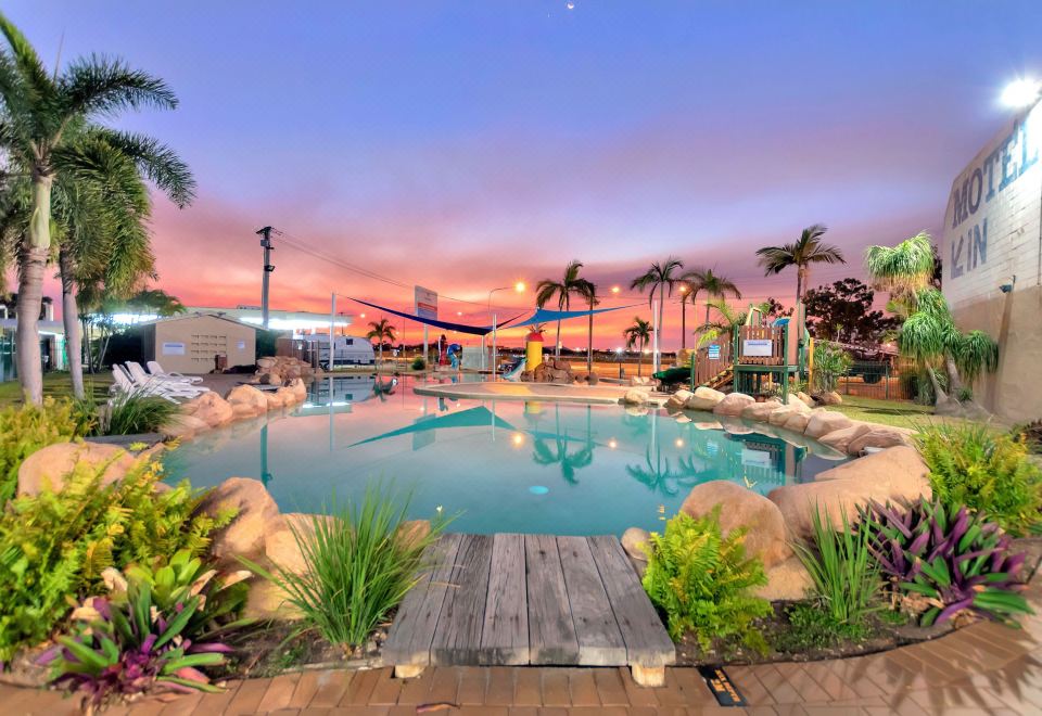 a large outdoor swimming pool surrounded by palm trees and buildings , with the sun setting in the background at Discovery Parks - Townsville