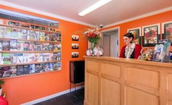 a receptionist standing behind a wooden counter in an orange - walled room , with shelves of books and vases on the walls at Victoria Lodge