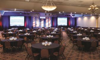 a large conference room with rows of tables and chairs , a stage at the end , and multiple screens on the walls at Bismarck Hotel and Conference Center