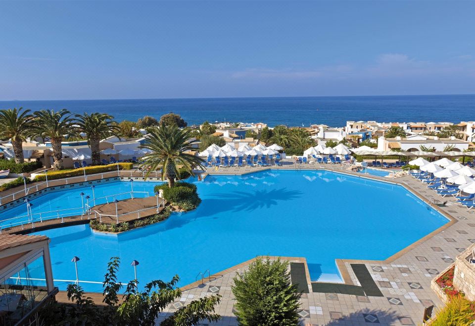 a large outdoor swimming pool surrounded by a resort , with a view of the ocean in the background at Aldemar Knossos Royal