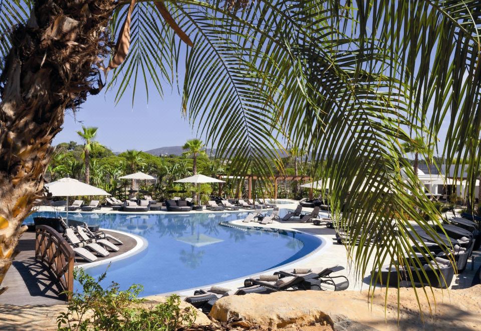 a large outdoor pool surrounded by palm trees , with lounge chairs and umbrellas placed around the pool area at Conrad Algarve