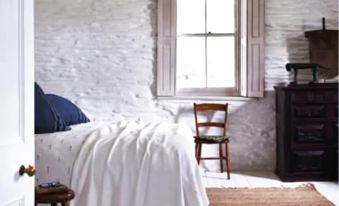 a bedroom with a white bed and brick wall , featuring a window with a wooden stool and a chair at The White House