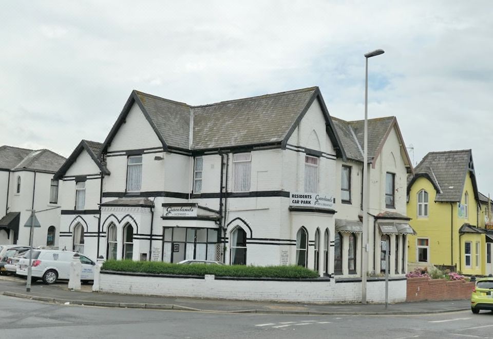 a two - story white building with black trim , situated in front of a brick building with a green lawn and a car parked in the street at Gracelands Guest House