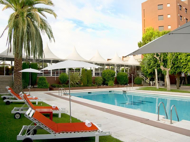 a large outdoor pool surrounded by lounge chairs and umbrellas , with palm trees in the background at Hotel America Igualada