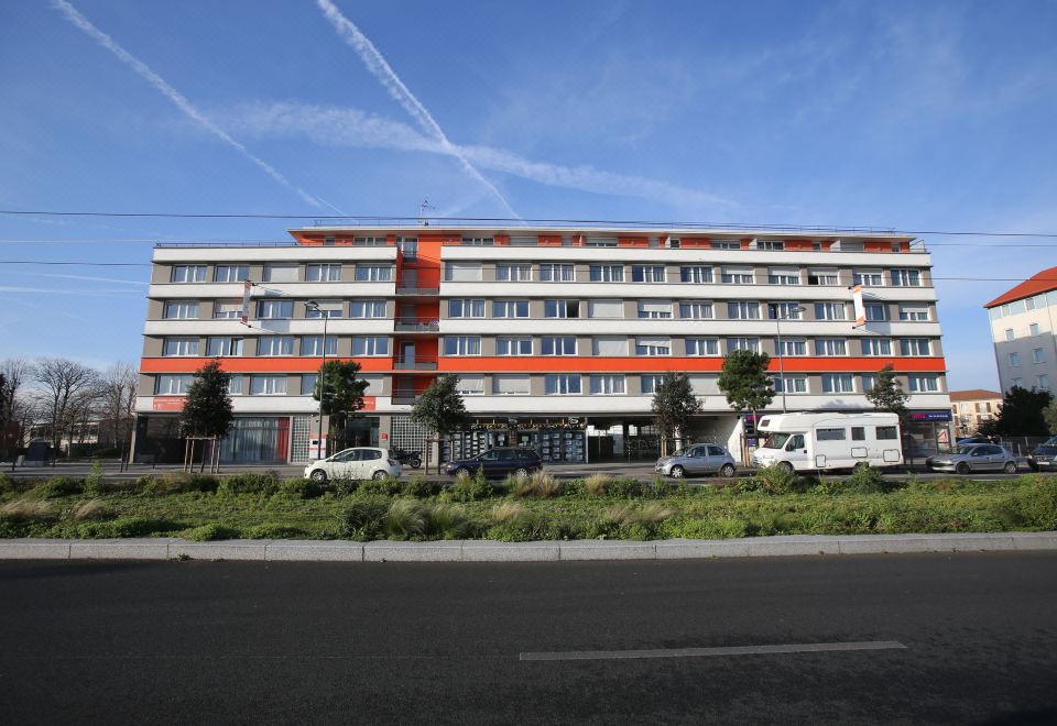 a large , multi - story building with a red and white facade , parked cars , and trees in the background at Adonis Paris Sud