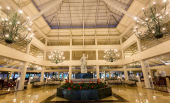 a large , white atrium with a statue of a man in the center , surrounded by chairs and tables at Bahia Principe Grand la Romana - All Inclusive