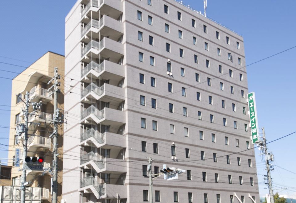 a tall , beige - colored apartment building with multiple floors and balconies , situated in front of other buildings and under a clear blue sky at Ace Inn Matsumoto