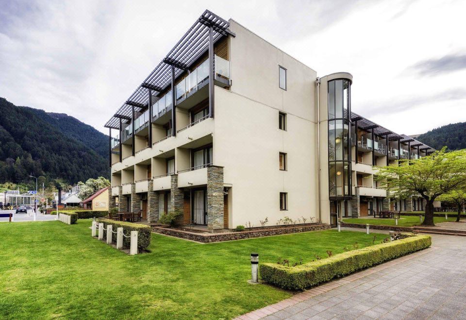 a modern apartment building surrounded by grass and trees , with a view of the mountains in the background at Novotel Queenstown Lakeside