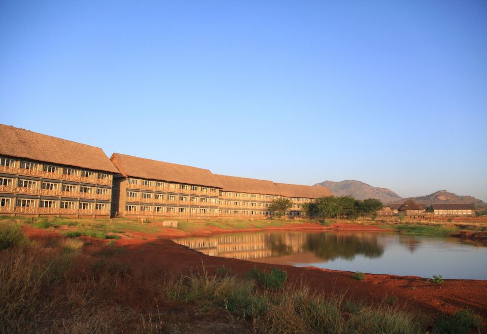 a large building with a red roof is reflected in the water , surrounded by trees and mountains at VOI Wildlife Lodge