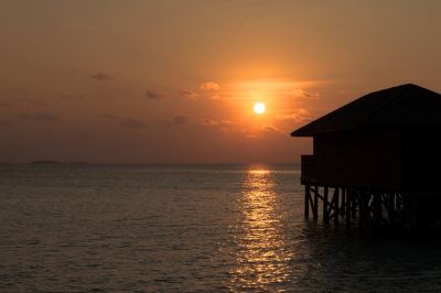 a sunset over the ocean with a small hut on stilts , and the sun setting in the background at Vilamendhoo Island Resort & Spa