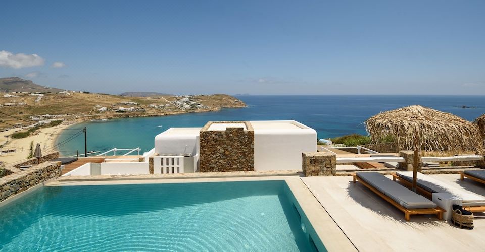 a large swimming pool is surrounded by a white structure with a stone pillar and an ocean view at The Summit of Mykonos