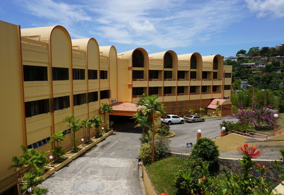 a large , beige - colored building with arched windows and balconies , surrounded by tropical plants and cars parked in a parking lot at Ambassador Hotel