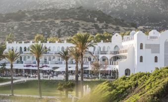 a large white building surrounded by a grassy field , with palm trees in the background at Falkensteiner Resort Capo Boi