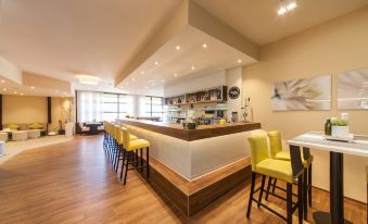 a modern kitchen with yellow chairs and a white counter , surrounded by wooden flooring and large windows at Select Hotel Erlangen