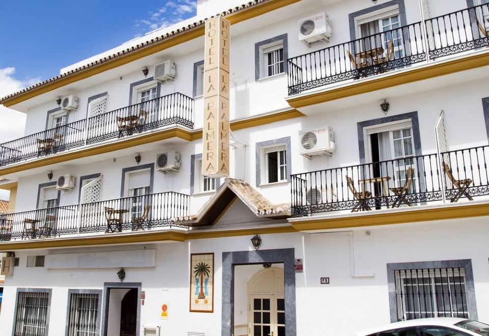 a two - story white building with balconies and a car parked in front , under a clear blue sky at La Palmera Rooms