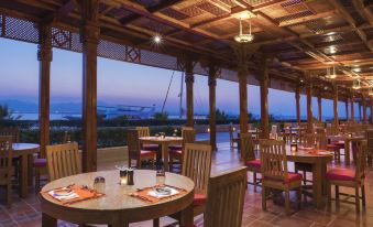 a restaurant with wooden tables and chairs , surrounded by a view of the ocean at dusk at Kempinski Hotel Soma Bay