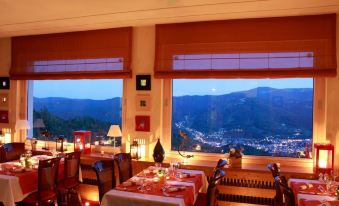 a restaurant with large windows offering a view of the city and mountains , featuring a dining table set for a meal at Hotel Panorama
