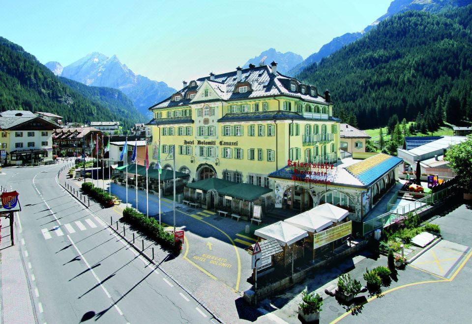 a large yellow building with a parking lot and mountains in the background , under a clear blue sky at Hotel Dolomiti Schloss