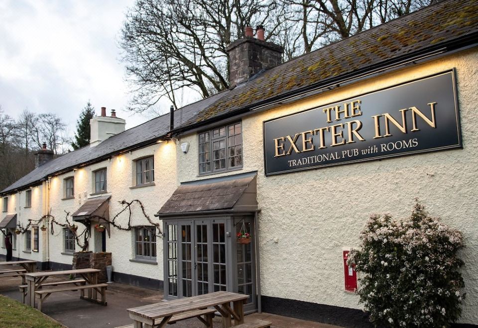 "the exterior of a small pub with white walls and black sign reading "" the exeter inn "" above the entrance , and picnic tables and benches outside" at The Exeter Inn