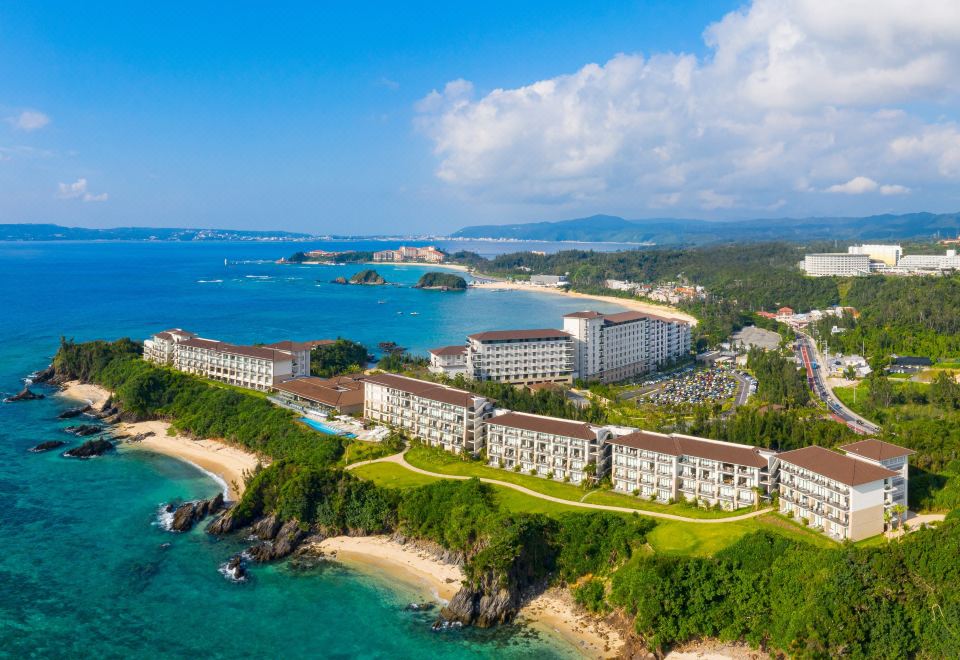 a large hotel is situated on a hillside overlooking the ocean , with a beach and mountains in the background at Halekulani Okinawa