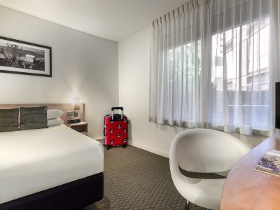 a hotel room with a bed , a red suitcase , and a chair near a window at Ibis Melbourne Hotel and Apartments