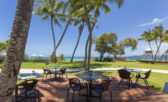 a tropical setting with palm trees , umbrellas , and tables under the shade of umbrellas on a wooden deck overlooking the ocean at Chateau Royal Beach Resort & Spa, Noumea