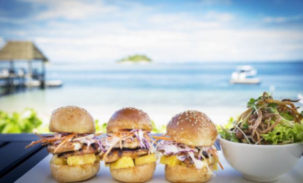 three grilled burgers with various toppings are served on a table near the ocean , and a bowl of salad is placed nearby at Malolo Island Resort