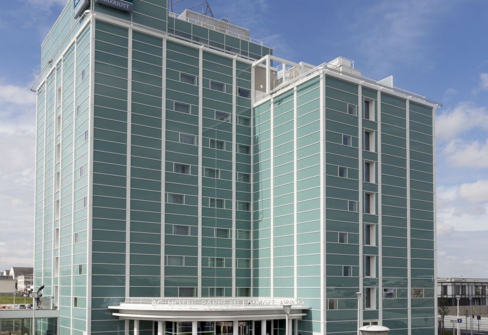 "a tall building with a green facade and the logo "" ac "" on top is surrounded by a parking lot" at AC Hotel Paris le Bourget Airport