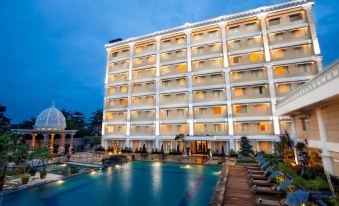 a large white hotel with multiple floors , lit up at night , and a swimming pool in front of it at The Rich Jogja Hotel