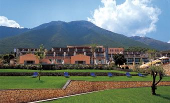 A large building with mountains in the background and green grass on either side is situated in an open area at Ionian Blue Bungalows and Spa Resort