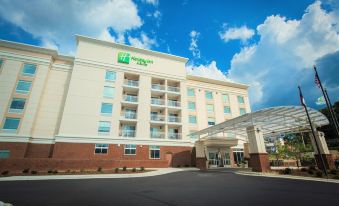 a large , modern hotel building with a green logo and a glass entrance , surrounded by trees and under a clear blue sky at Holiday Inn & Suites Arden - Asheville Airport