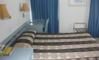 a small bedroom with a blue bed , a desk , and a chair , along with a window and curtains at Bridge Motel